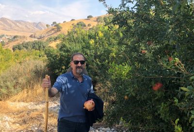 Jaffer holding a hiking stick in one hand and a fruit that looks like a mango in the other. He is wearing sunglasses and he is standing in front of trees, grasslands, and mountains.