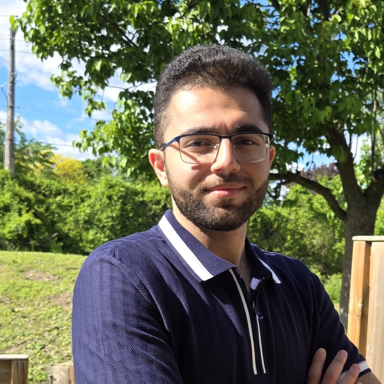 Omar with arms folded standing in front of a field and some green trees.