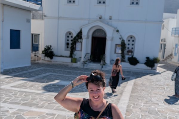 Janett removes her sunglasses in front of a white building in Greece with blue turrets and flying a Greek flag.