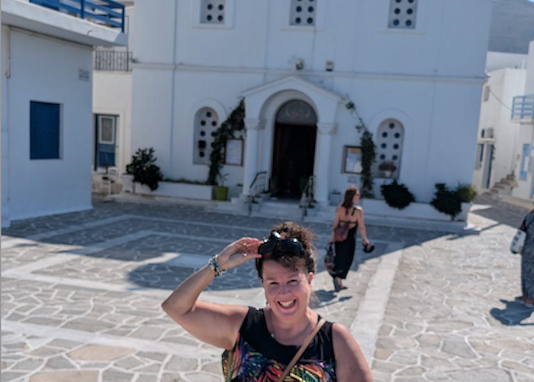 Janett removes her sunglasses in front of a white building in Greece with blue turrets and flying a Greek flag.