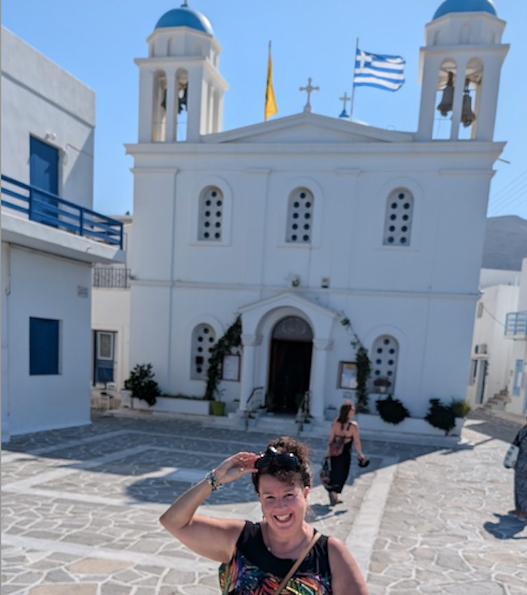 Janett removes her sunglasses in front of a white building in Greece with blue turrets and flying a Greek flag.