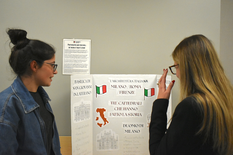 Gabriella shows her poster to Rebecca. Hand drawn sketches of three famous cathedrals in pencil.
