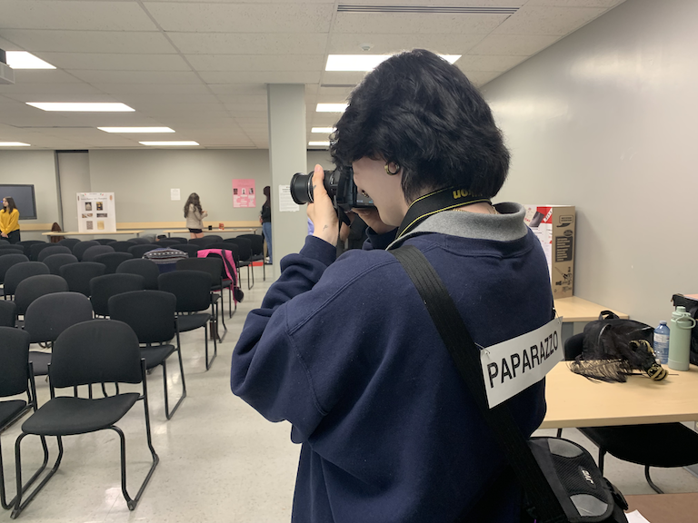 Student with a Paparazzo sign on her back takes photos of the audience.