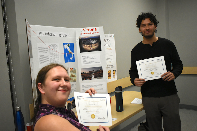 Aurora and Timothy show off their certificates in front of their poster about anfiteatri.