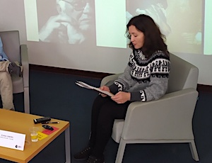 Ioana sitting in a chair reading from her paper. In the background book covers are projected on a wall.