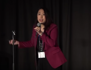 Ai in red jacket stands at mic on stage at LSA