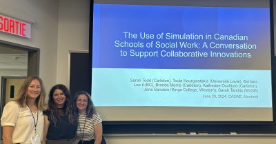 Jane Saunders, Sarah Tarshis, and Katherine Occhiuto posing in front of a large screen with the title of their conversation circle "The Use of Simulation in Canadian Schools of Social Work: A Conversation to Support Collaborative Innovation"