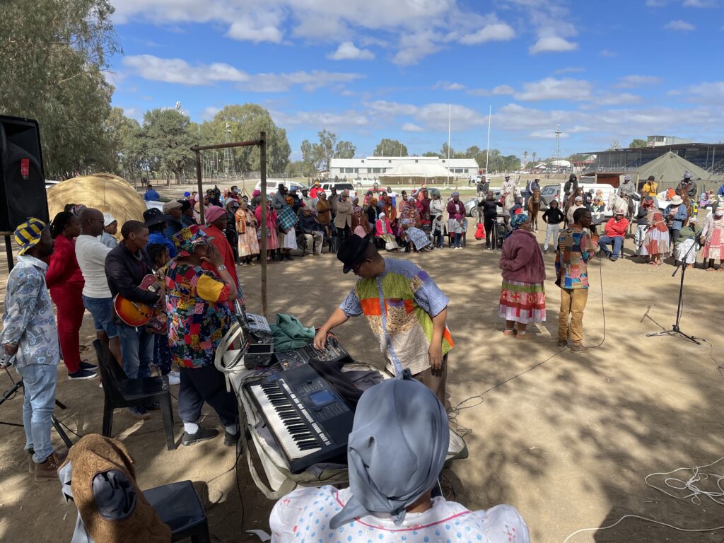 A crowd gathered outdoors with a traditional Nama hut in the background