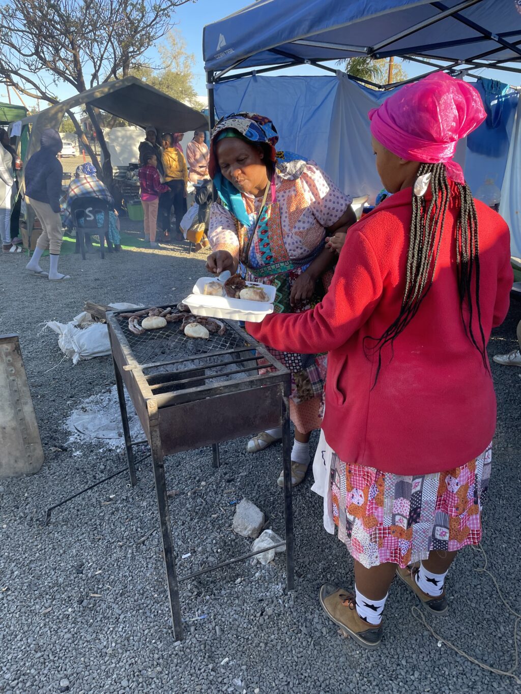 Two vendors dressed in traditional Nama clothing preparing a meal of meat and bread.