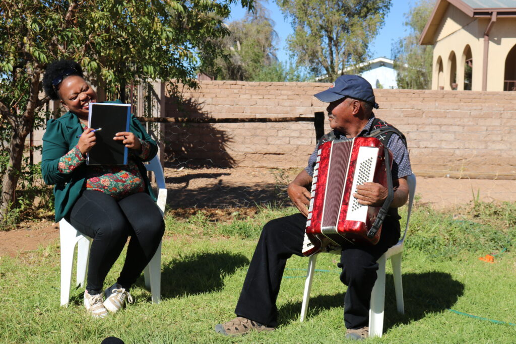 Dr. Sharon Gabie & Mr. Andries Snyders sitting on white chairs, holding a notebook and an accordion respectively.