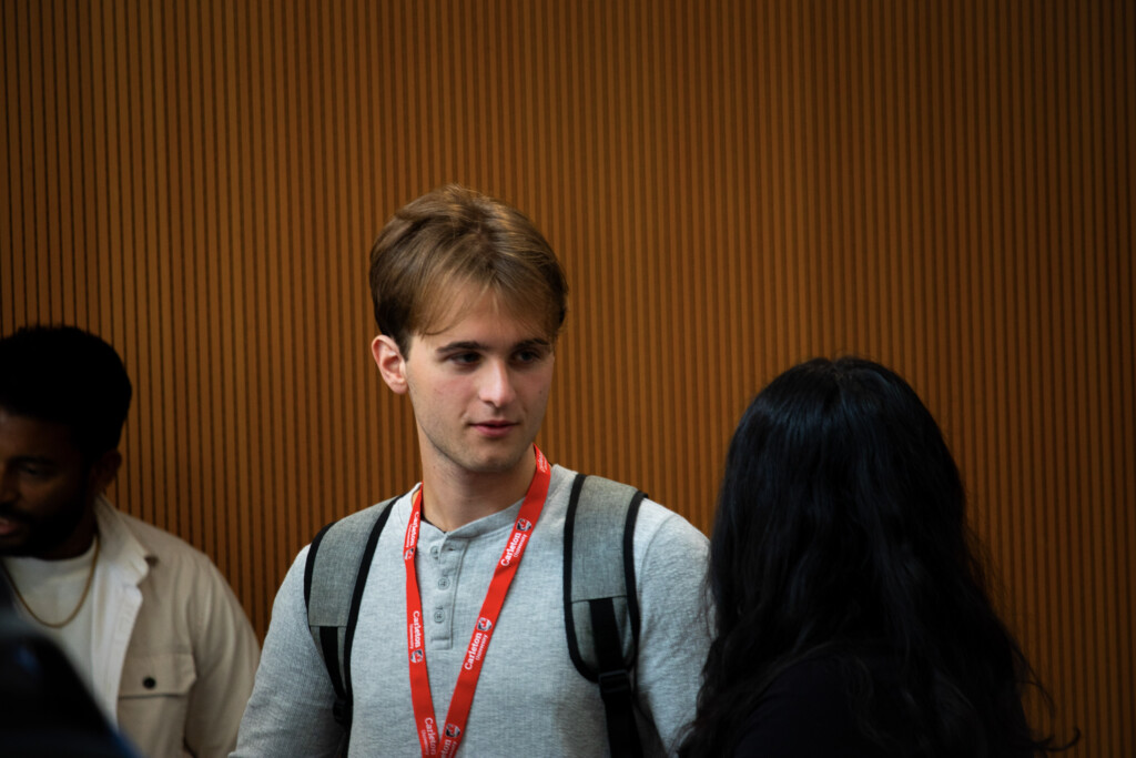 Two attendees wearing Carleton lanyards talk during the Alumni Reception in front of a wood-paneled wall.