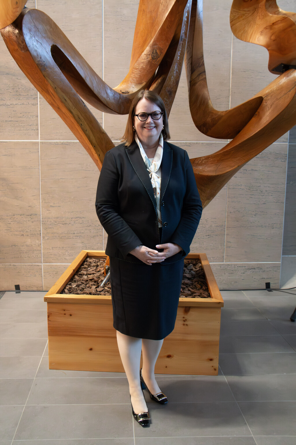 [name] poses near in front of a wooden sculpture.