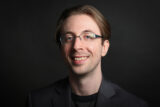 Photo of Benjamin Faveri, smiling with brown long hair and black glasses