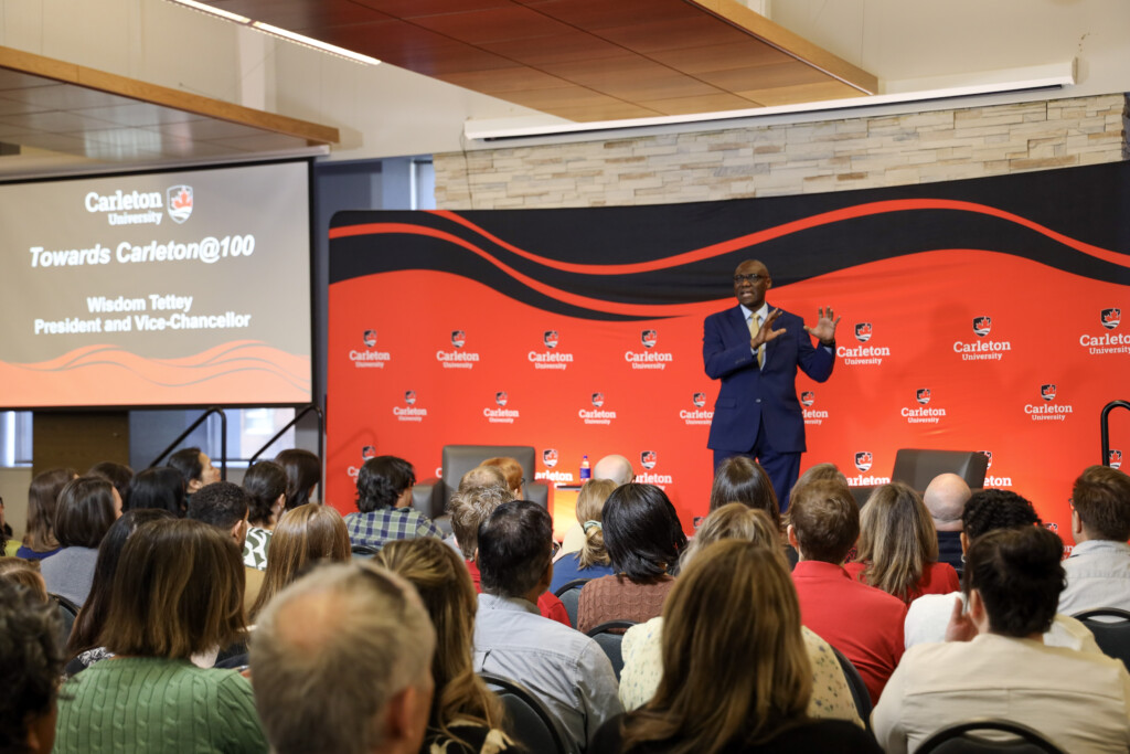 Room of people looking at a man standing on stage, pointing to a PowerPoint slide.