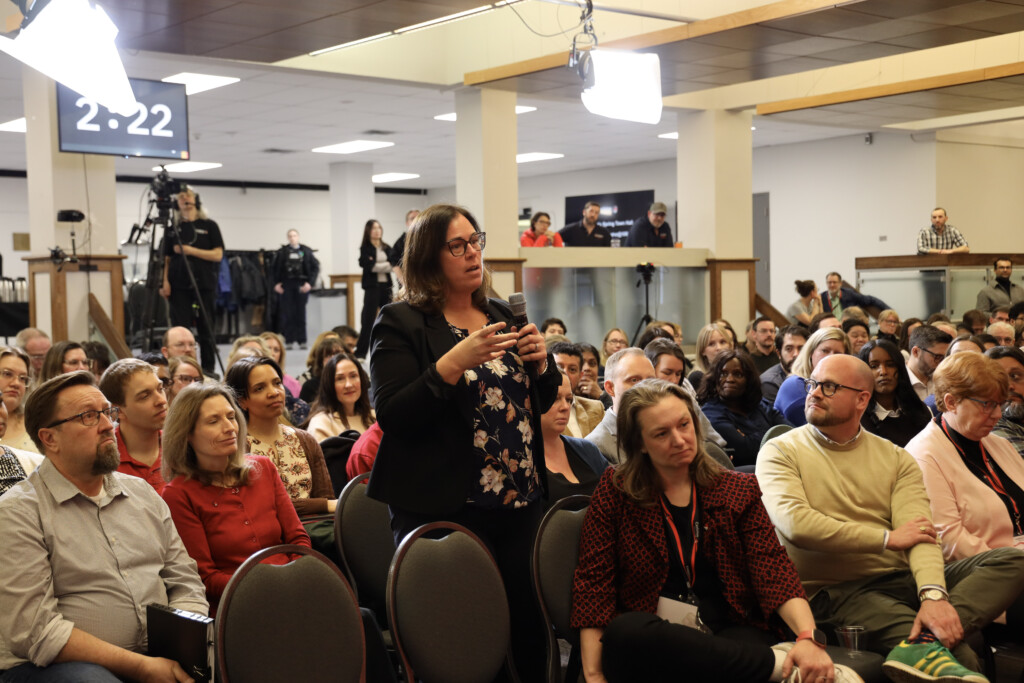 A room of people looking at a woman in the center holding a microphone.