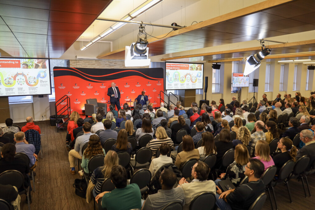 Room of people looking at a man standing on stage, pointing to a PowerPoint slide.