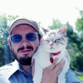 Colin Killby proudly holds his beautiful white and grey cat. Colin wears sunglasses and a ballcap, the cat has closed eyes and a pink collar with a red tag.