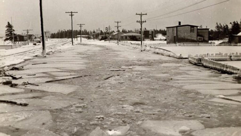 a black and white photograph showing a village with a flood of deep, icy water coursing along a main street.