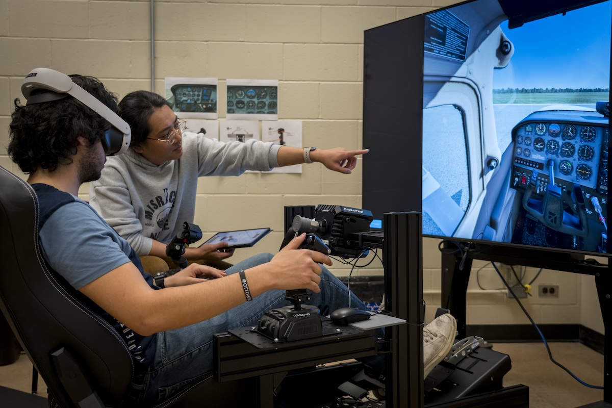 One student sits in the flight simulator as the pilot, while another student, who is the copilot, points at the screen.