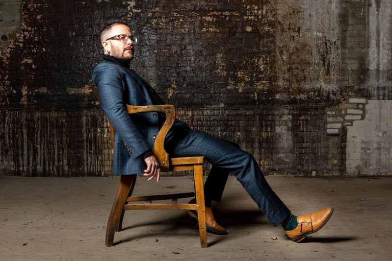 Cáel M. Keegan, a white man with short hair and glasses, leans back in a wooden chair in front of a distressed brick wall. He is wearing a sharp blue suit and light brown dress shoes.