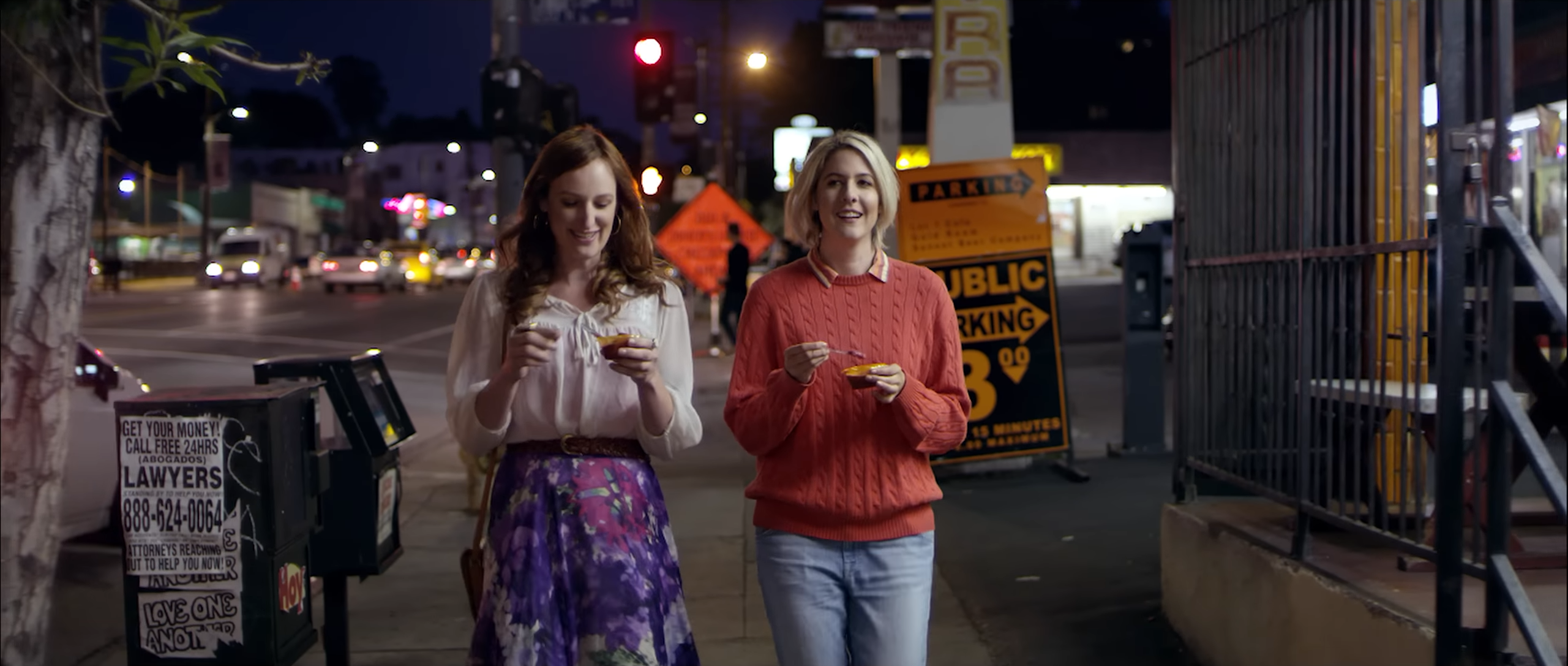 A city sidewalk at night. Two women walk toward the camera. On the left is a white trans woman who grins while looking down at her ice cream. She is wearing a loose white blouse and pink and purple floral long skirt. On the right a white cis woman with bleached blonde straight hair to her chin, a red cotton sweater and light-colored jeans. She talks as she holds her ice cream and spoon in her each hand.