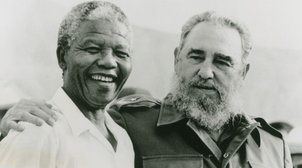 An older black man in a white shirt and with grey hair is embraced by a man in military fatigues, sporting a large grey beard