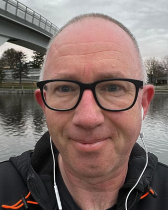 An aging, balding white man with glasses outside, with a bridge and a canal in the background.