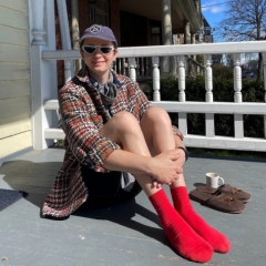 Me, a white person in my 30s, sitting on a sunny porch. Outfit includes: Anti-Stairs Club lounge hat, triangle sunnies, a thick houndstooth button down, bare legs, bright red socks. Next to me are my dollar store slippers and a tiny cup of coffee. Wide smile for Yo-Yo who is taking the photo.