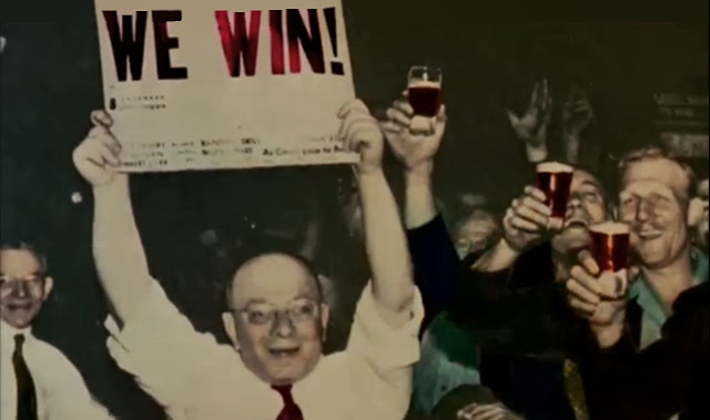 A man holds aloft a newspaper which declares "We win!" while other men raise their drinks in celebration.