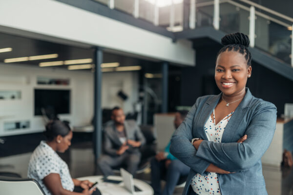 A woman standing with her arms crossed and smiling. In a office with people in the background talking to one another.