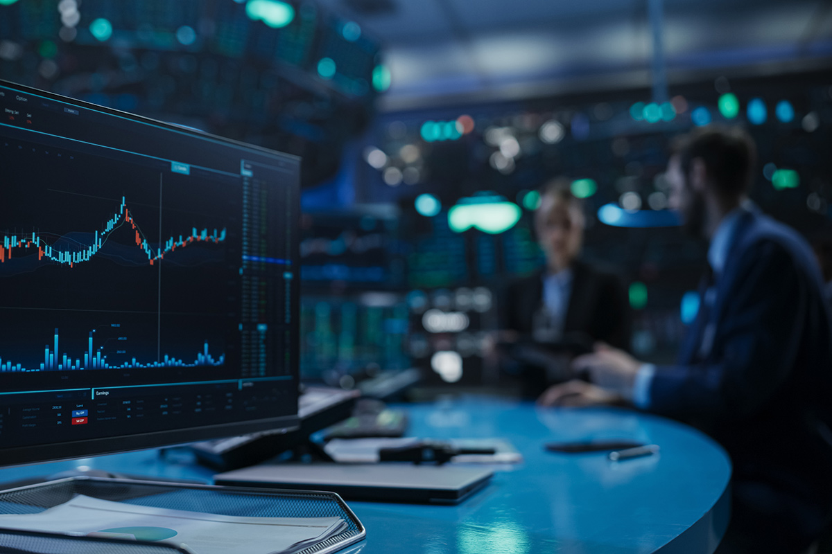 A computer monitor displays a stock market chart, with two people visible in the background.