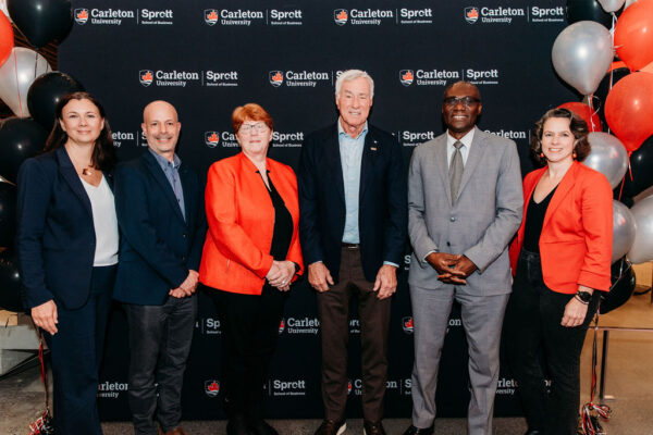 A group of six people smiling and posing in front of a step and repeat banner with the Sprott School of Business logo. There are balloons to the right-side of the image.